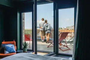 A couple stand on a balcony and look out of a window.