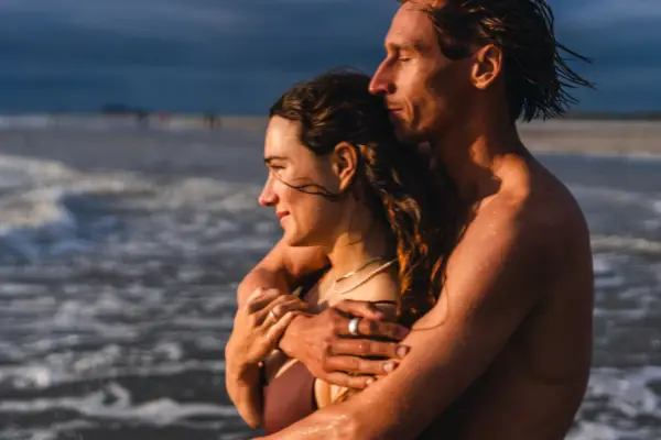 Ein Mann umarmt eine Frau am Strand und beide schauen auf das Meer.