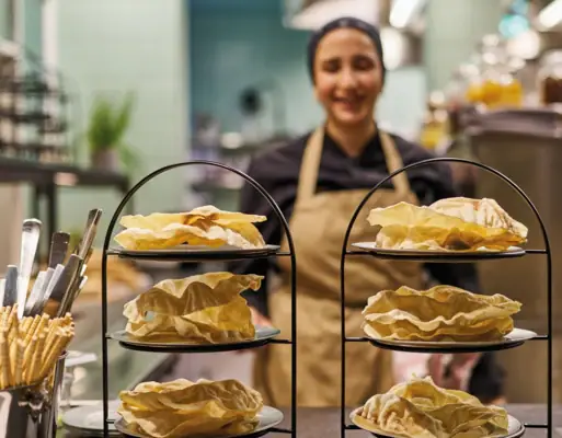 Two tiered trays stand in a kitchen and a cook stands behind them wearing an apron.