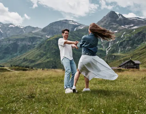 A man and woman dancing in a grassy field with mountains in the background.