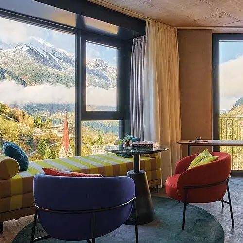 Modern eingerichtetes Hotelzimmer mit Panoramafenstern und Ausblick auf verschneite Alpen, Kirchturm und das Tal von Bad Gastein