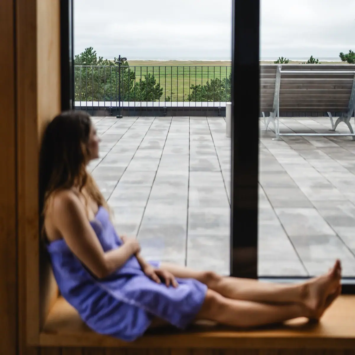 A woman is sitting on a windowsill in the sauna at the Urban Nature Hotel in St. Peter-Ording, looking out of the window.