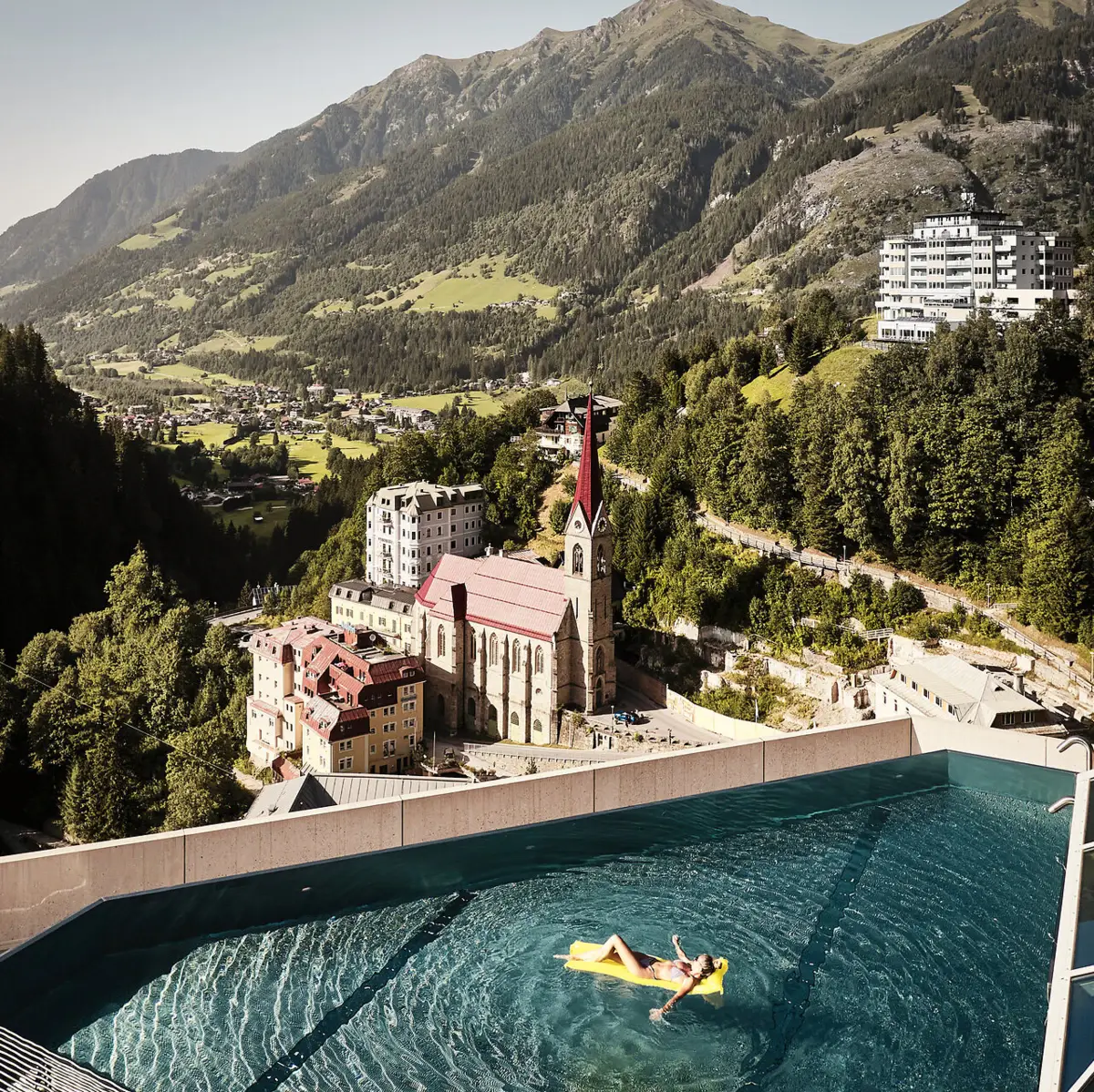 Infinity pool with a view of Bad Gastein and the Alpine landscape - woman relaxing on a yellow air mattress with a view of the church and historic buildings