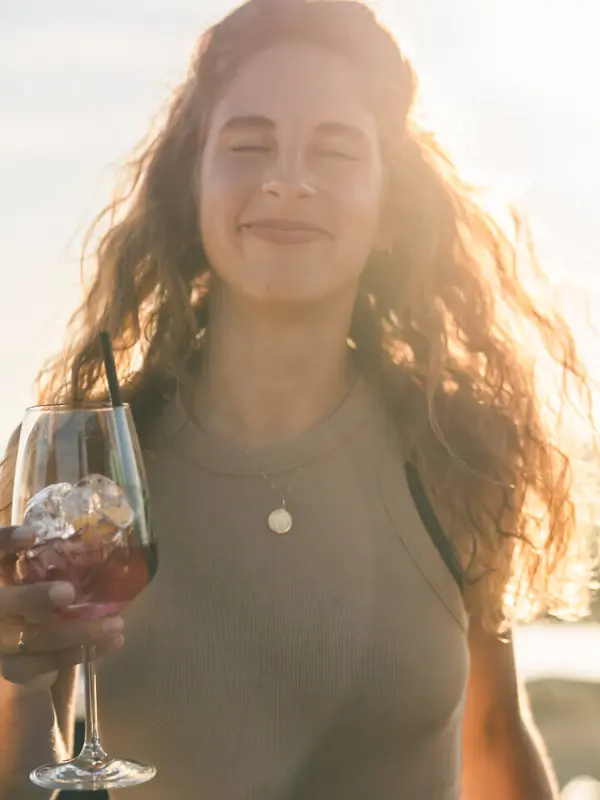 A woman smiles and holds a drink in a wine glass in her hand, with the sun shining behind her.