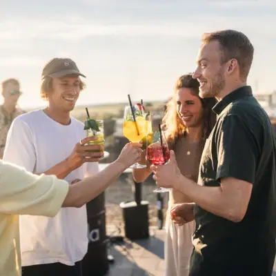 A group of people stand on the rooftop bar at Scraper's Club St. Peter-Ording with drinks in hand and a view of the salt marshes and the sea.