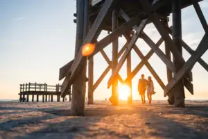 A couple walks on the beach under a wooden structure.