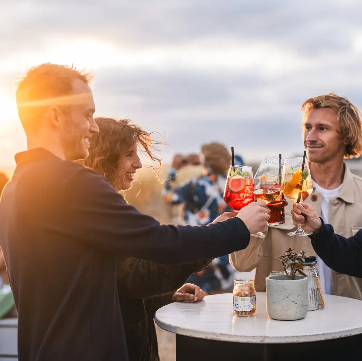 A group of people are standing on the rooftop bar Scraper's Club St. Peter-Ording with drinks in their hands, toasting the sunset.