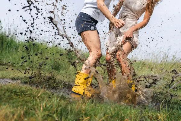 Zwei Frauen springen mit gelben Gummistiefeln in eine Matschpfütze.