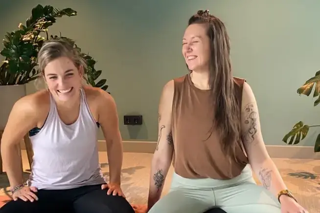 Meike & Agnes Two women sit cross-legged on a yoga mat, smiling.