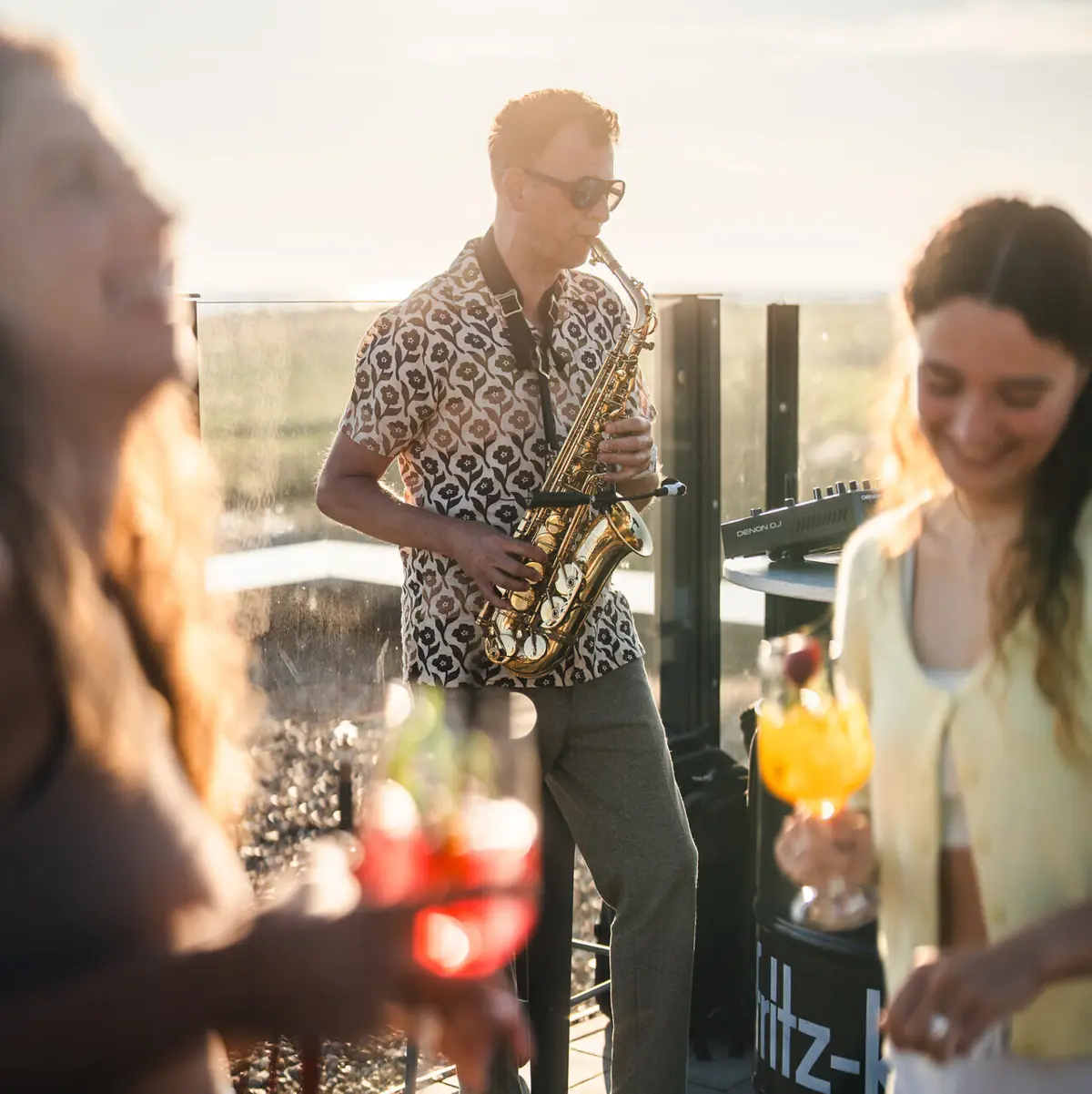 A saxophonist plays on a rooftop terrace while people dance with drinks in their hands.