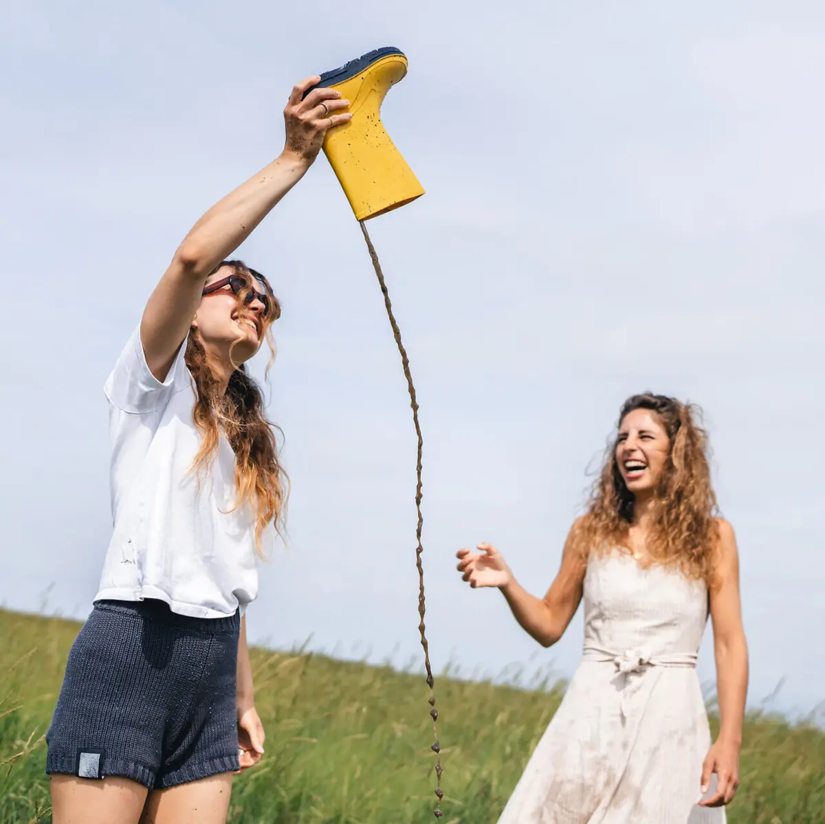 A woman is emptying a rubber wellington boot, whilst another woman stands next to her, laughing.