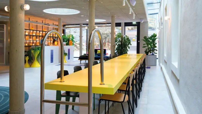 Kreatives Interior im Hotel:Modern gestalteter Rezeptionsbereich mit Schwimmbadleiter als Designelement, gelb-weißen Akzenten und stilvollen Sitzgelegenheiten.