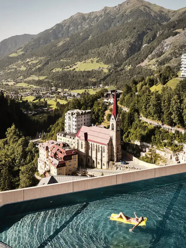 Infinity Pool mit Ausblick auf Bad Gastein und die Alpenlandschaft – Frau entspannt auf gelber Luftmatratze mit Blick auf Kirche und historische Gebäude