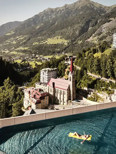 Infinity pool with a view of Bad Gastein and the Alpine landscape - woman relaxing on a yellow air mattress with a view of the church and historic buildings