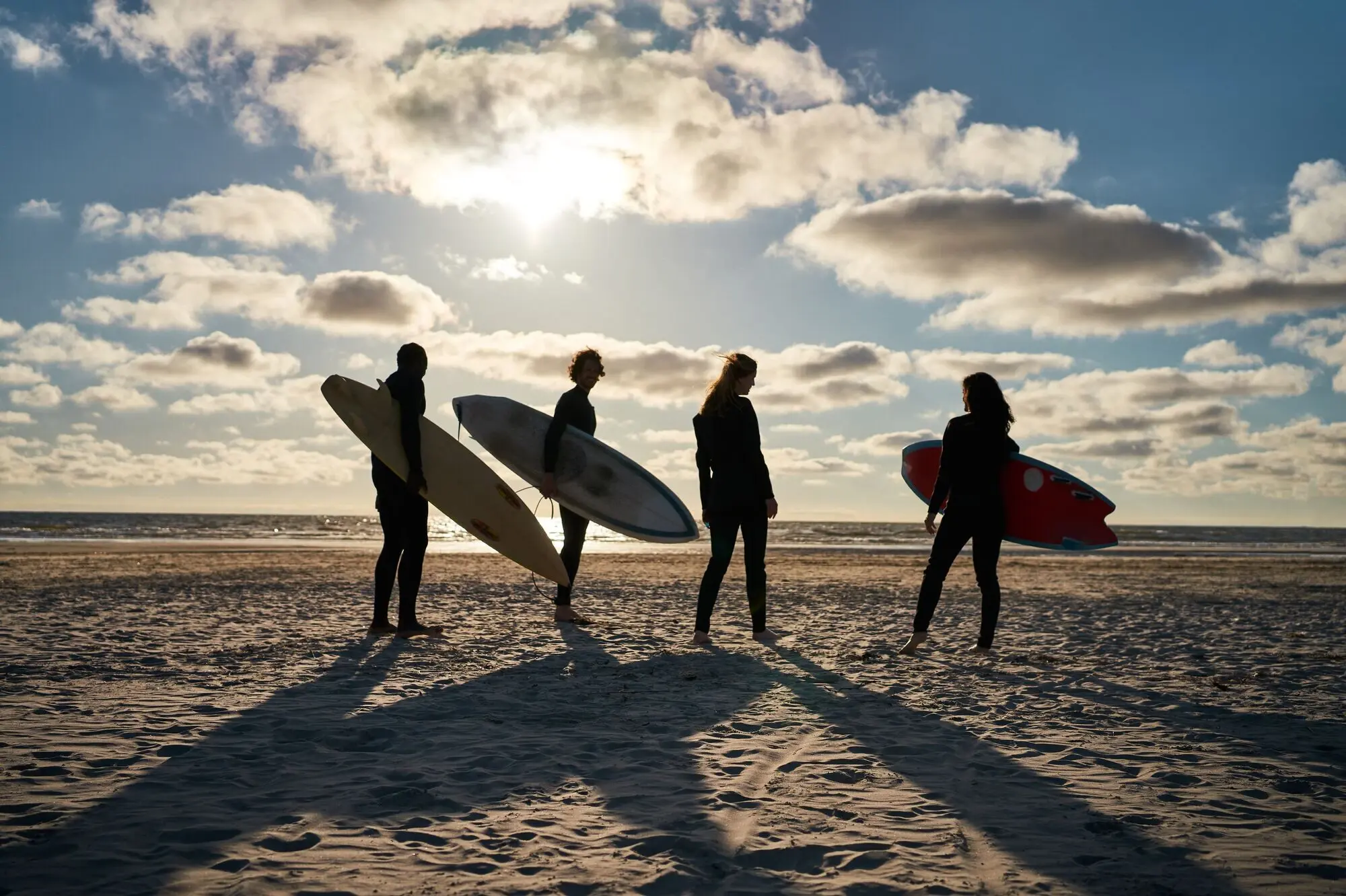 Vier Menschen stehen an einem Strand und tragen Surfbretter unter dem Arm bei Sonnenschein.