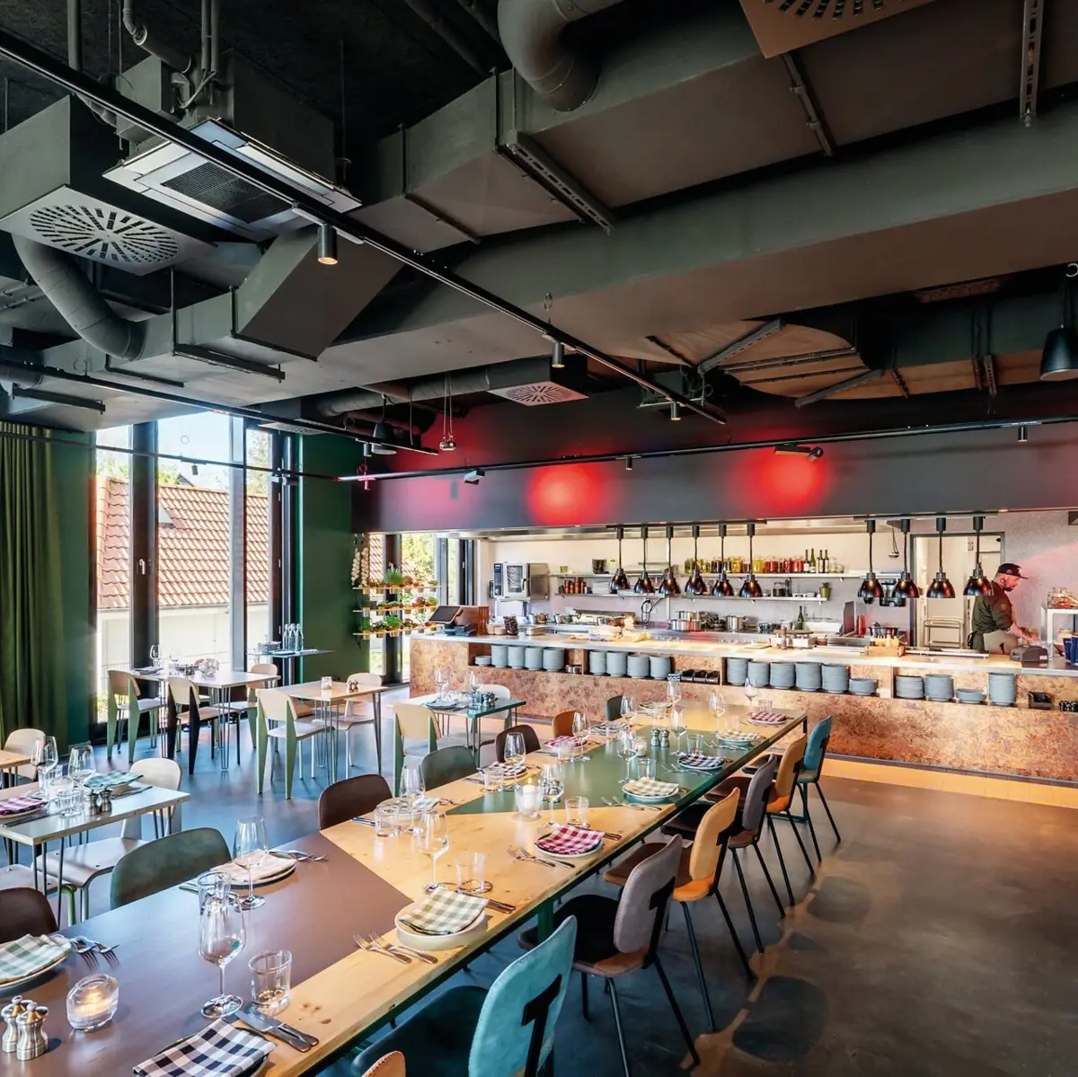 A restaurant with a long table and several set tables and an open kitchen in the background.