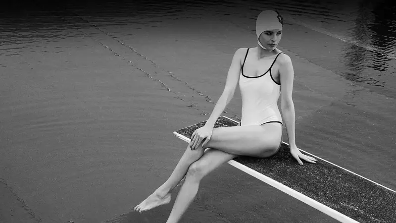 A woman in a swimsuit and swimming cap sits on a diving board.