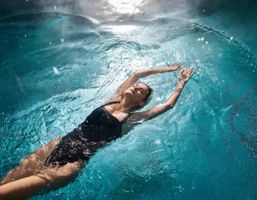 Alpine Swim Club A woman in a black swimsuit is floating peacefully on her back in a sun-drenched swimming pool.