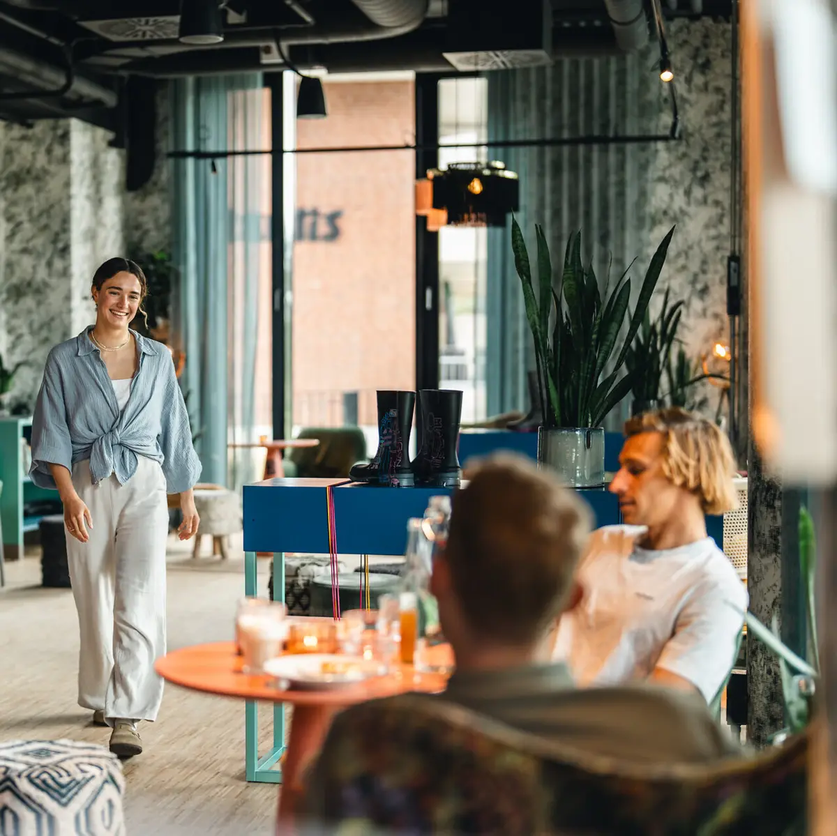 A woman walks up to a table in a bar at the Urban Nature Hotel where two men are already sitting and smiles.