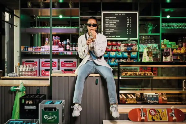 Eine junge Frau mit Sonnenbrille sitzt auf einer Theke in einem Kiosk nd trinkt aus einem Getränk mit Strohhalm.