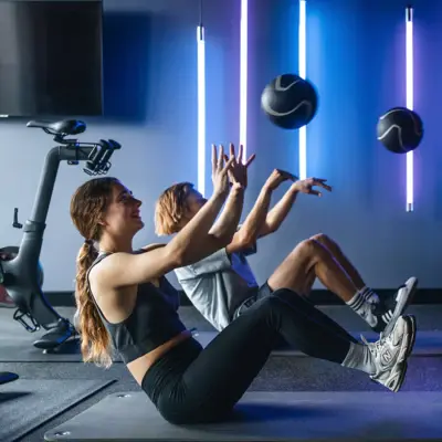 Two people are sitting on a mat in the gym, holding their legs and upper bodies in the air while throwing a ball.