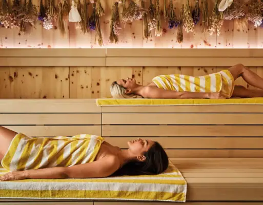 Two women with yellow and white striped towels lie relaxed in a modern sauna with wooden walls and dried flowers on the ceiling.