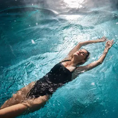 Frau in schwarzem Badeanzug treibt entspannt auf dem Rücken in einem sonnendurchfluteten Pool.