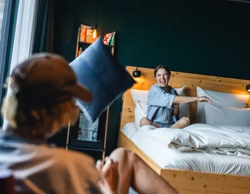 Woman sitting on a bed in an Urban Nature Hotel St. Peter-Ording room, playfully tossing a pillow toward the camera.