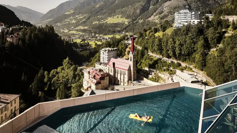 Infinity Pool mit Ausblick auf Bad Gastein und die Alpenlandschaft – Frau entspannt auf gelber Luftmatratze mit Blick auf Kirche und historische Gebäude