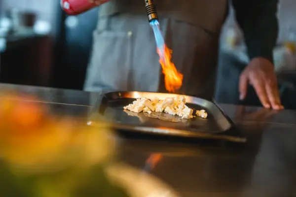 A chef flambés cauliflower with a small flamethrower in a kitchen.
