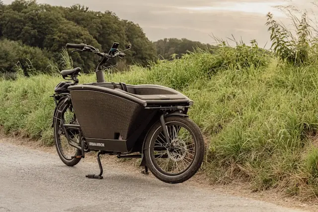 Ein Lastenfahrrad steht am Rand einer Straße neben einem grünen Wall und mit Bäumen im Hintergrund.