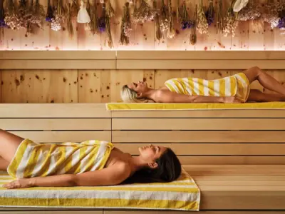Two women with yellow and white striped towels lie relaxed in a modern sauna with wooden walls and dried flowers on the ceiling.
