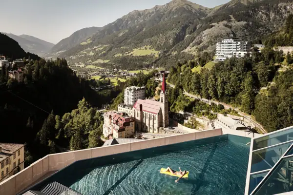 Rooftop Infinity Pool im Badeschloss Bad Gastein Infinity Pool mit Ausblick auf Bad Gastein und die Alpenlandschaft – Frau entspannt auf gelber Luftmatratze mit Blick auf Kirche und historische Gebäude