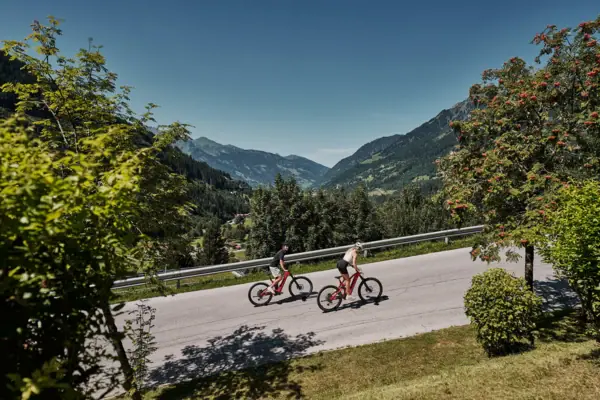 Summer in Bad Gastein Two people are cycling along a road; trees and mountains can be seen in the background.