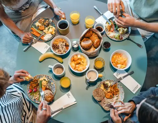 A round breakfast table photographed from above, with four people sitting around it eating bread rolls, fruit and other food.