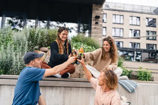 A group of people are sitting in the square outside the Urban Nature Hotel in St Peter-Ording, laughing and clinking their drink bottles together.