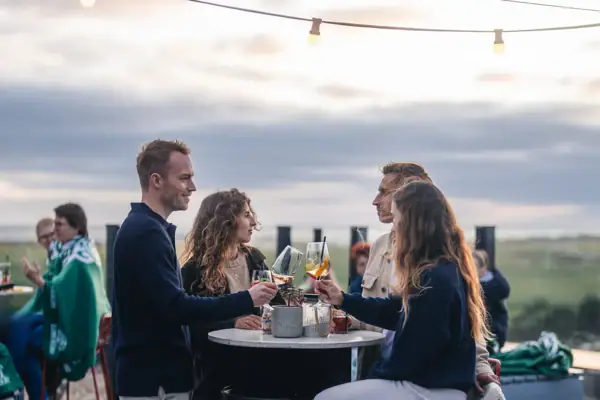 Eine Gruppe von Menschen steht auf der Rooftop Bar Scraper's Club St. Peter-Ording mit Getränken in der Hand und mit Blick auf die Salzwiesen und das Meer.