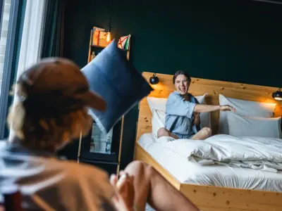 Woman sitting on a bed in an Urban Nature Hotel St. Peter-Ording room, playfully tossing a pillow toward the camera.